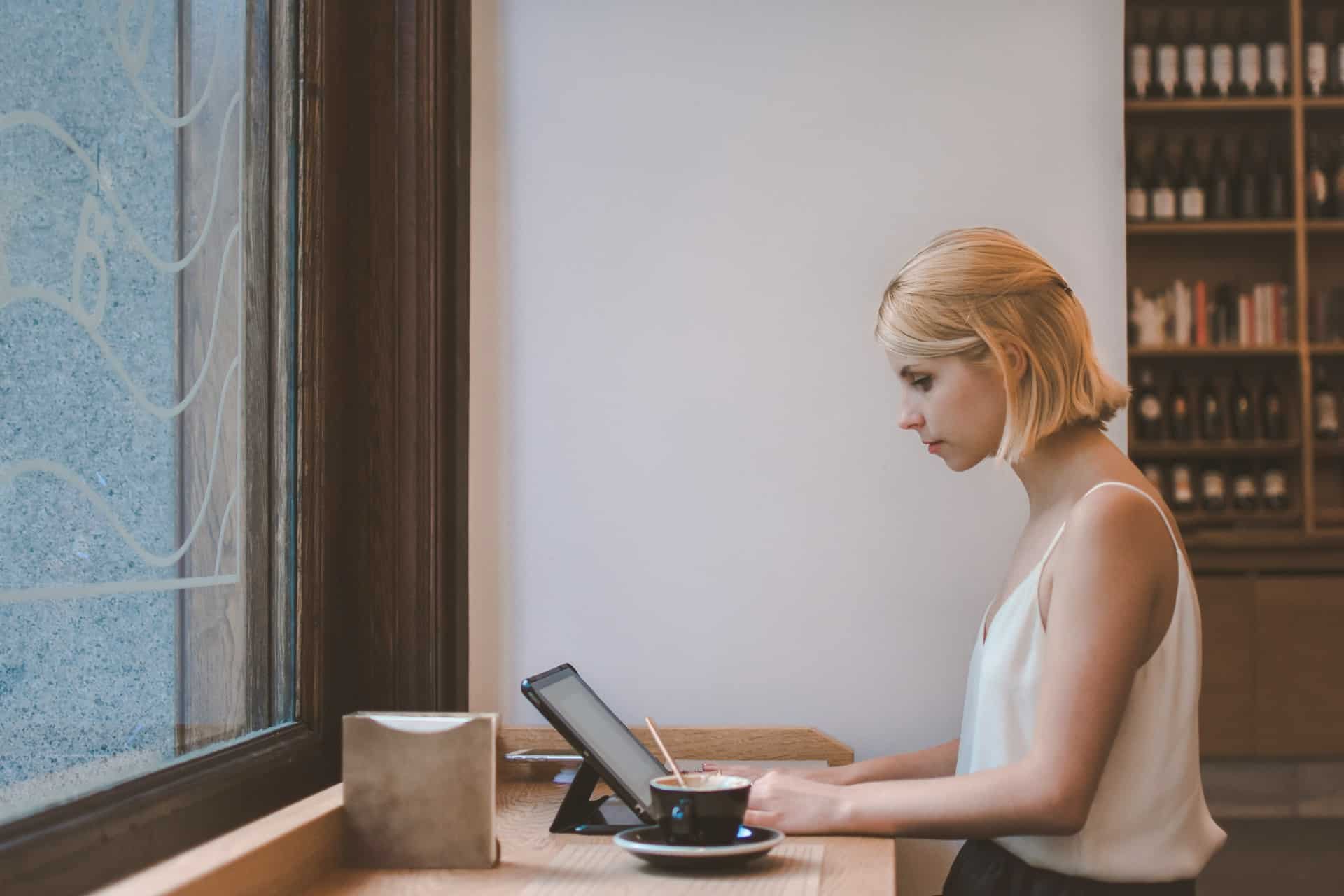 Woman in a white top working at a laptop in a cafe with a coffee beside her